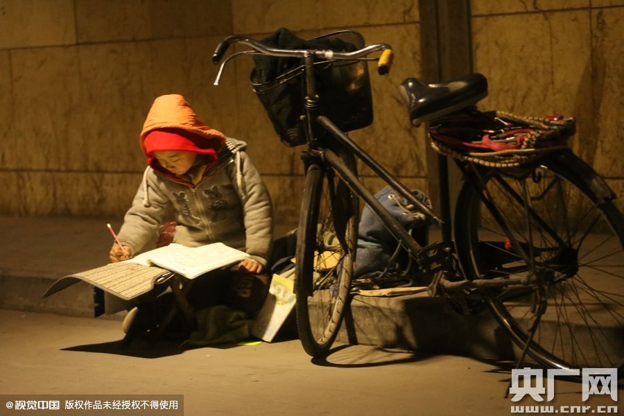 鄭州中原路京廣鐵路大橋下涵洞的馬路牙子邊，一位戴著紅色辣椒帽的小男孩趴在一個(gè)小馬扎上，上面鋪著一張車墊，在微弱的燈光下寫作業(yè)